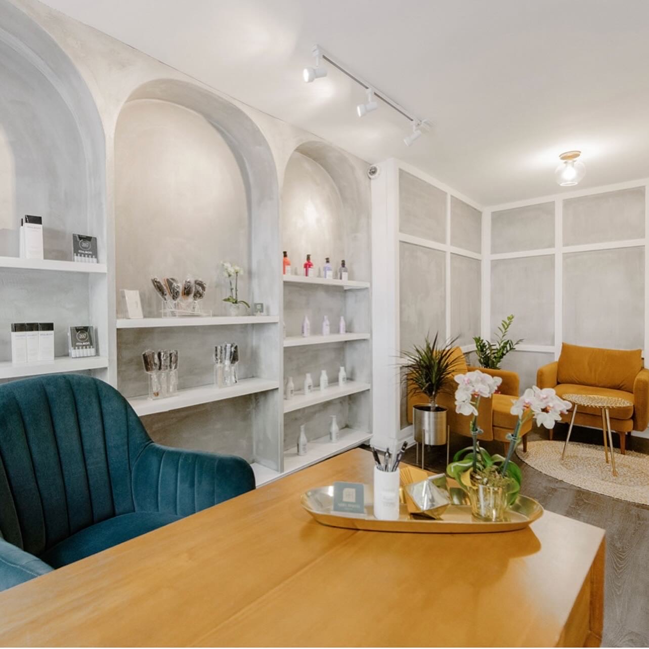 Modern salon interior with blue chair, wooden table, display shelves, and mustard armchairs with plants.